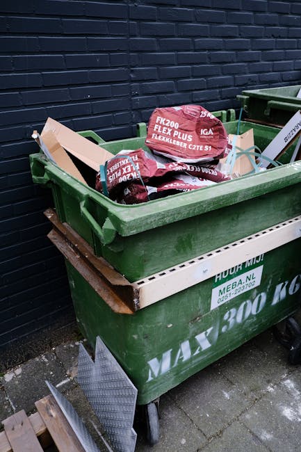 An outdoor scene showing two large green waste bins positioned on a paved surface beside a dark brick wall. The bins are filled with various discarded items, including several bags of garden or waste 