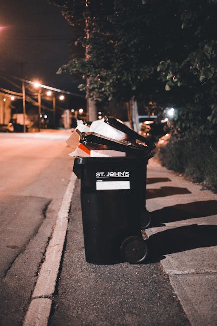 An outdoor scene showing two large green waste bins positioned on a paved surface beside a dark brick wall. The bins are filled with various discarded items, including several bags of garden or waste 