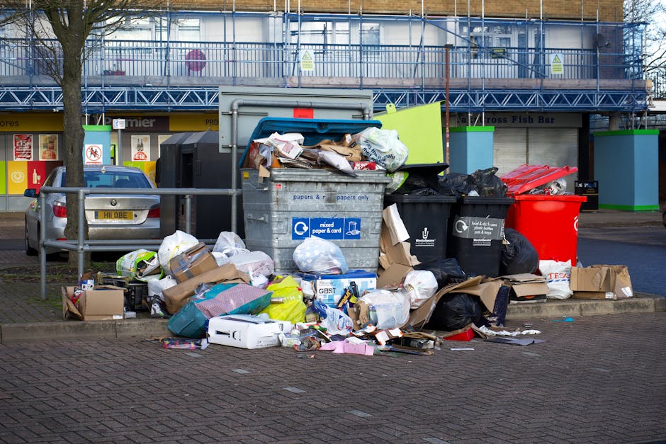 A large collection of mixed waste and rubbish bags, including black, green, and red plastic bags, cardboard boxes, and various discarded packaging, piled on a paved sidewalk near a metal railing and a